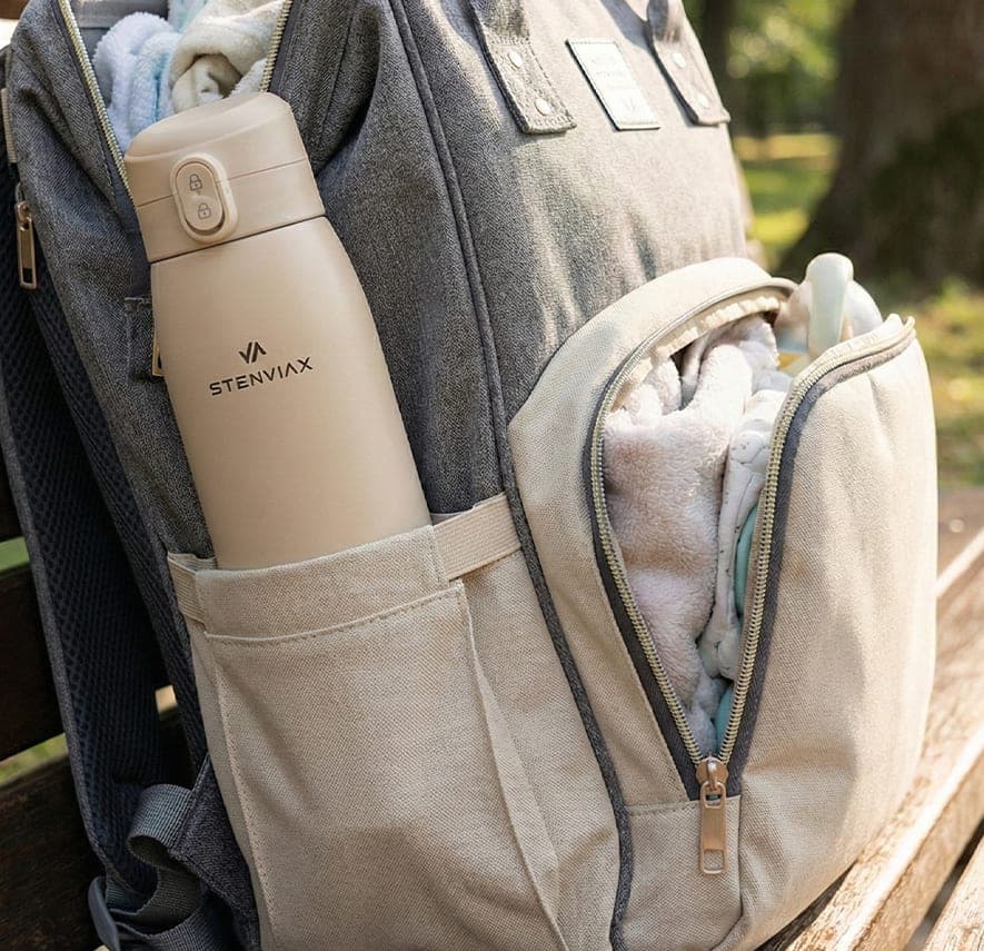 The Silk Beige warmer standing in the side pocket of a changing backpack on a park bench