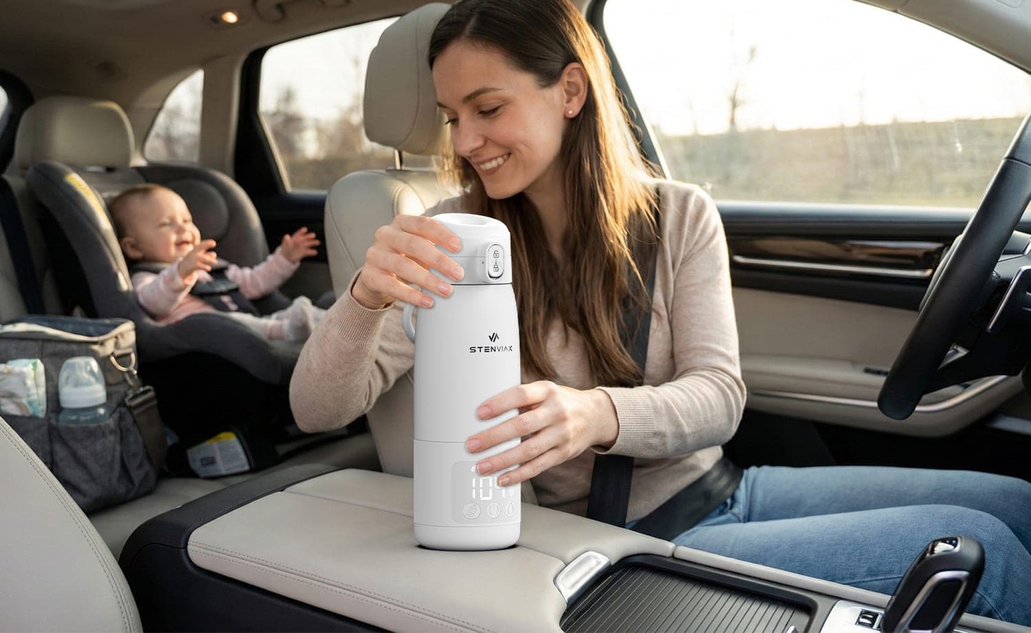A smiling parent in the driver's seat holding the Luxe White warmer on the center console, a baby laughing in a car seat behind