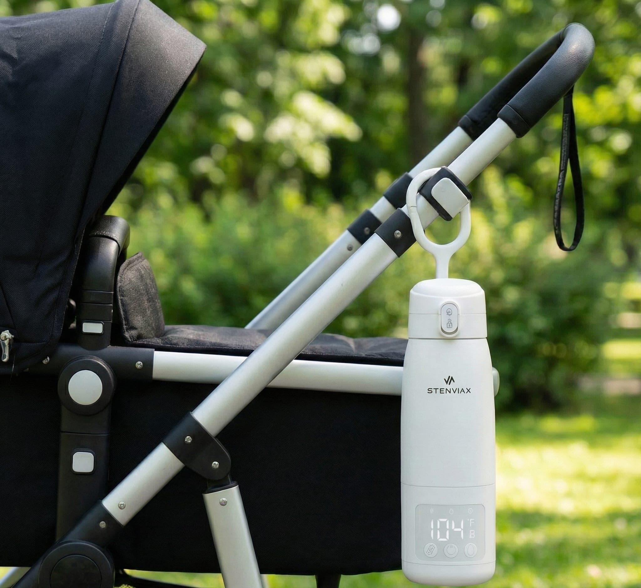 The Luxe White warmer clipped by its handle to a stroller frame in a park, its display reading 104°F